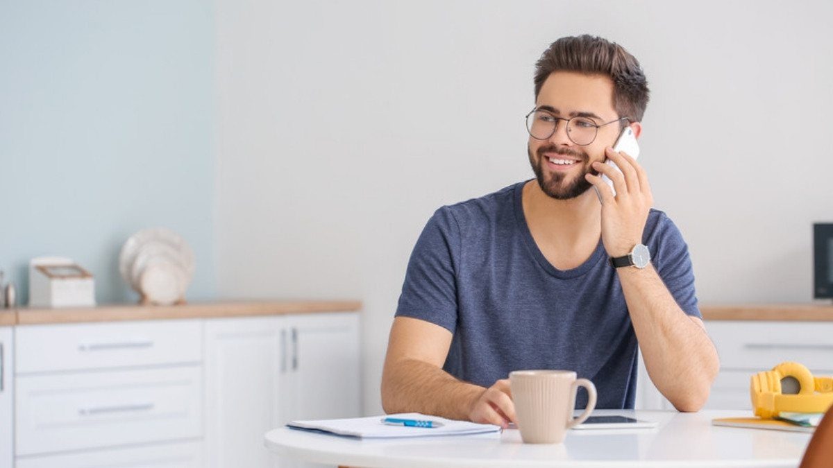 A young man talks on his phone at a bright kitchen table, a coffee mug and an open notepad close at hand