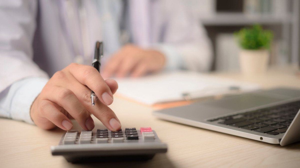 A hand taps figures into a calculator beside an open laptop while totaling medical bills at a desk