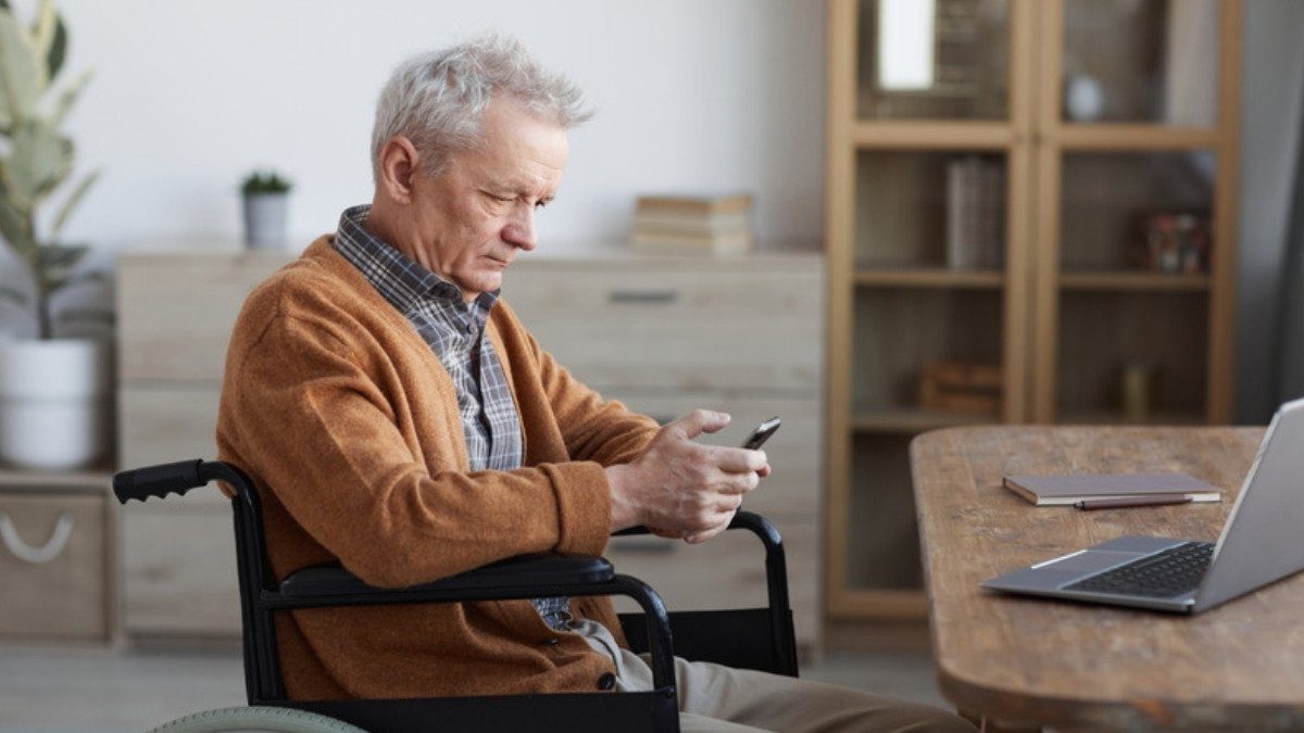 A gray haired man in a wheelchair checks his phone in a sunny living room, getting ready to make a call