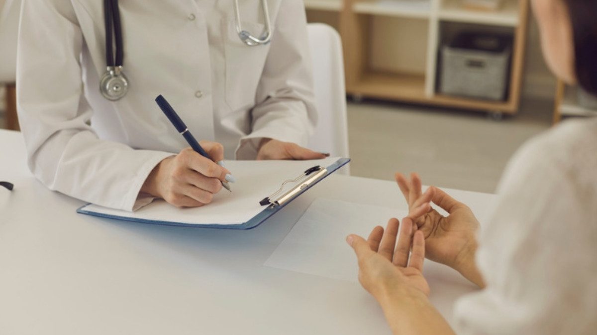 A doctor in a white coat writes on a clipboard while a patient sits across the desk.