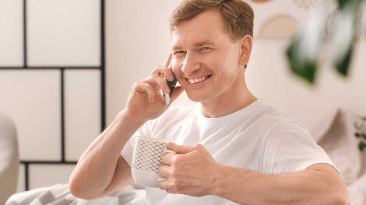A smiling man in a white shirt talks on his phone and holds a mug while sitting at home on a bright morning