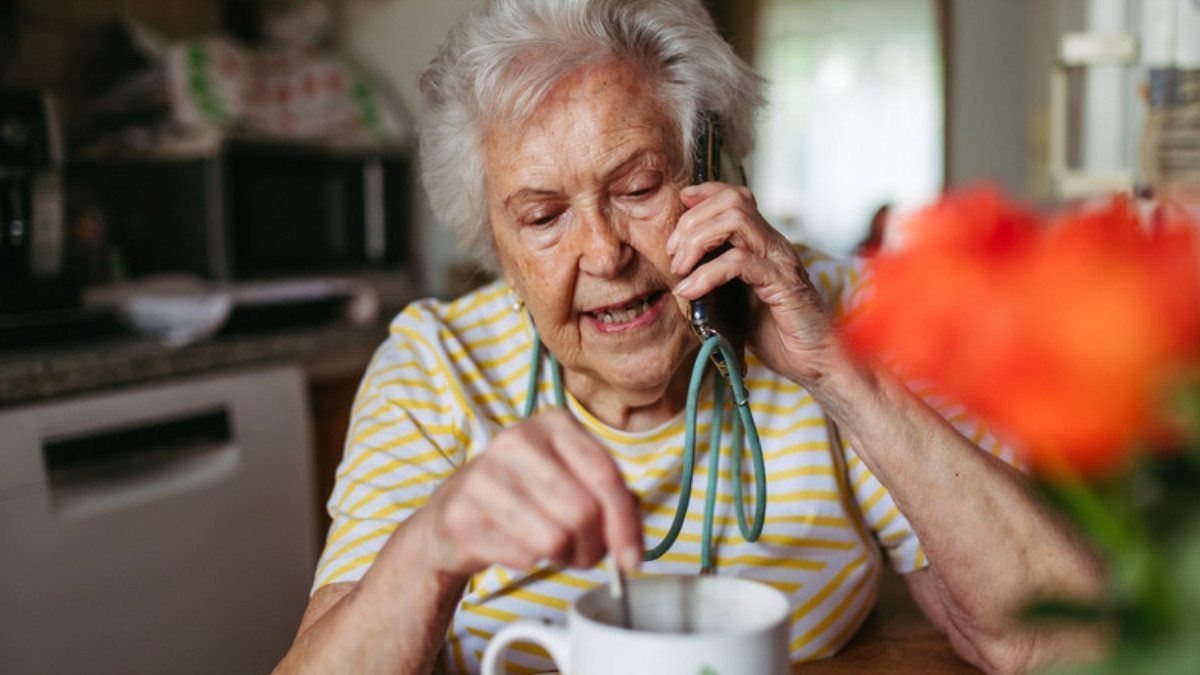 An older woman stirs her tea while talking on the phone at home, asking questions about a late injury claim