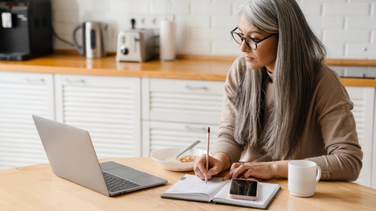 A woman writes notes at her kitchen table beside a laptop and phone, listing the bills and pay stubs in her car accident case