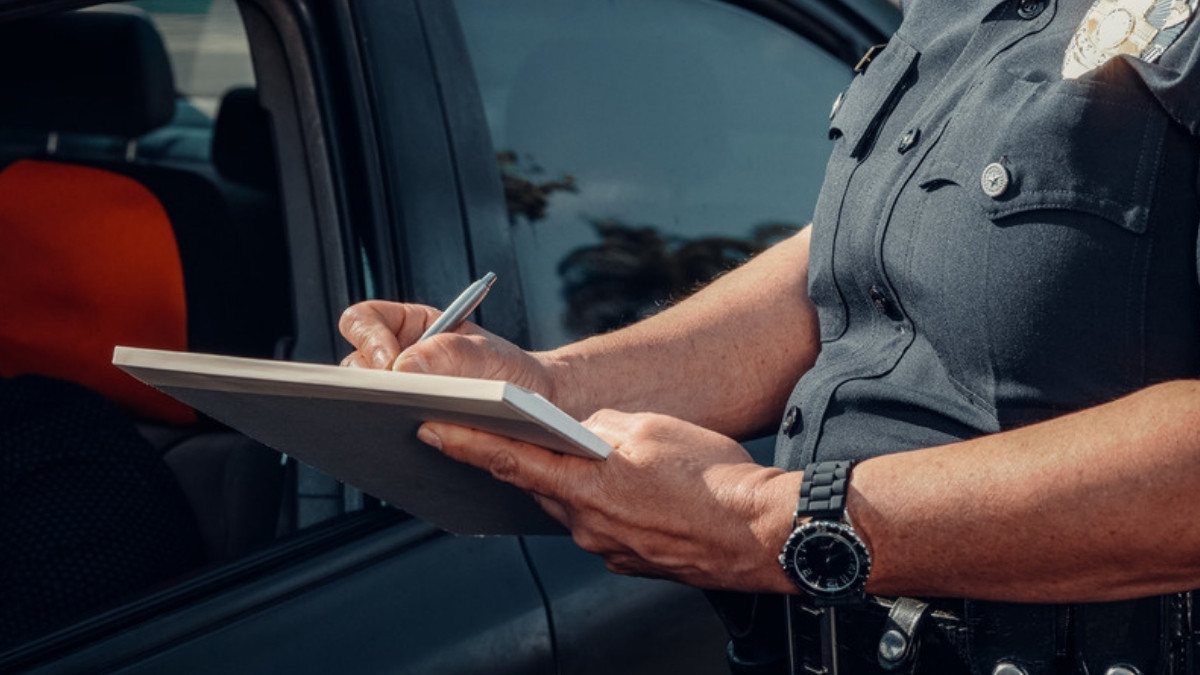 An officer writes on a clipboard beside a dark car, noting details that will later appear in the accident report