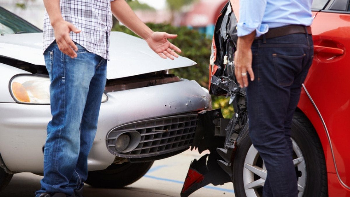 Two drivers stand between a silver car and a red car with crushed fronts, arguing over which one was at fault