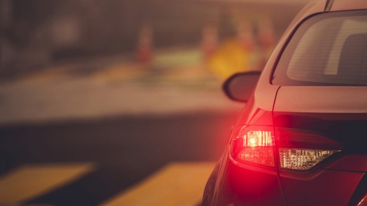 A red car waits at a painted crosswalk with its rear signal lamp glowing as the driver gets ready to turn