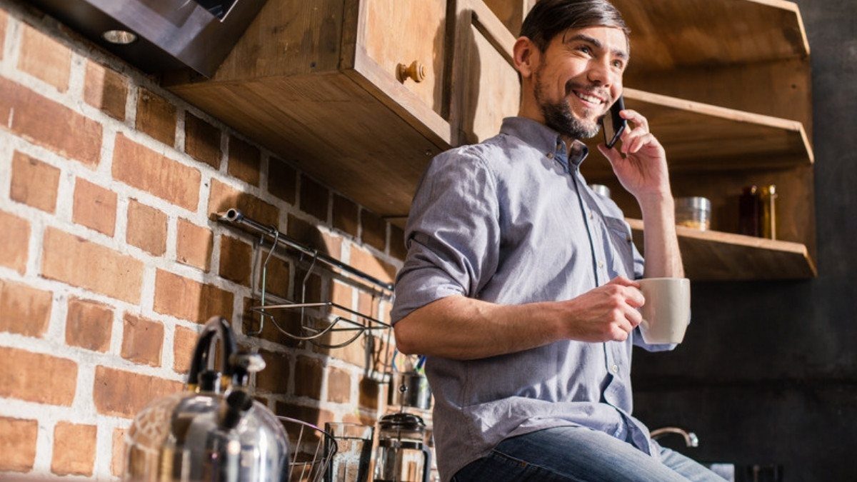 A man perched in his brick walled kitchen holds a coffee mug and smiles as he talks on his phone
