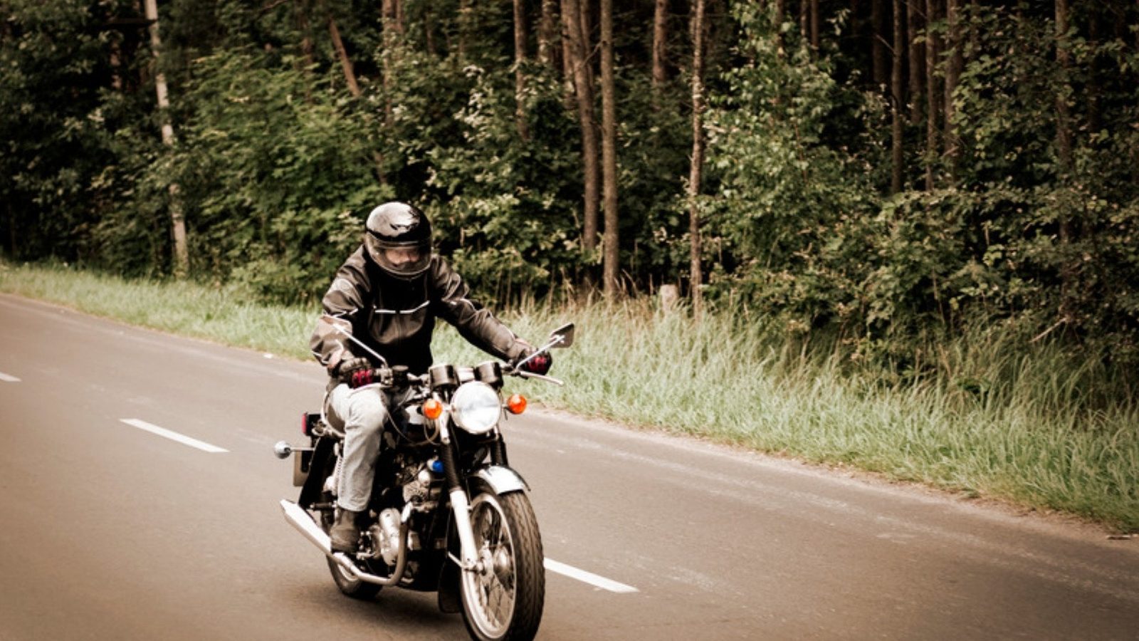 A rider in a full face helmet and leather jacket rides a motorcycle down a two lane road past tall trees.