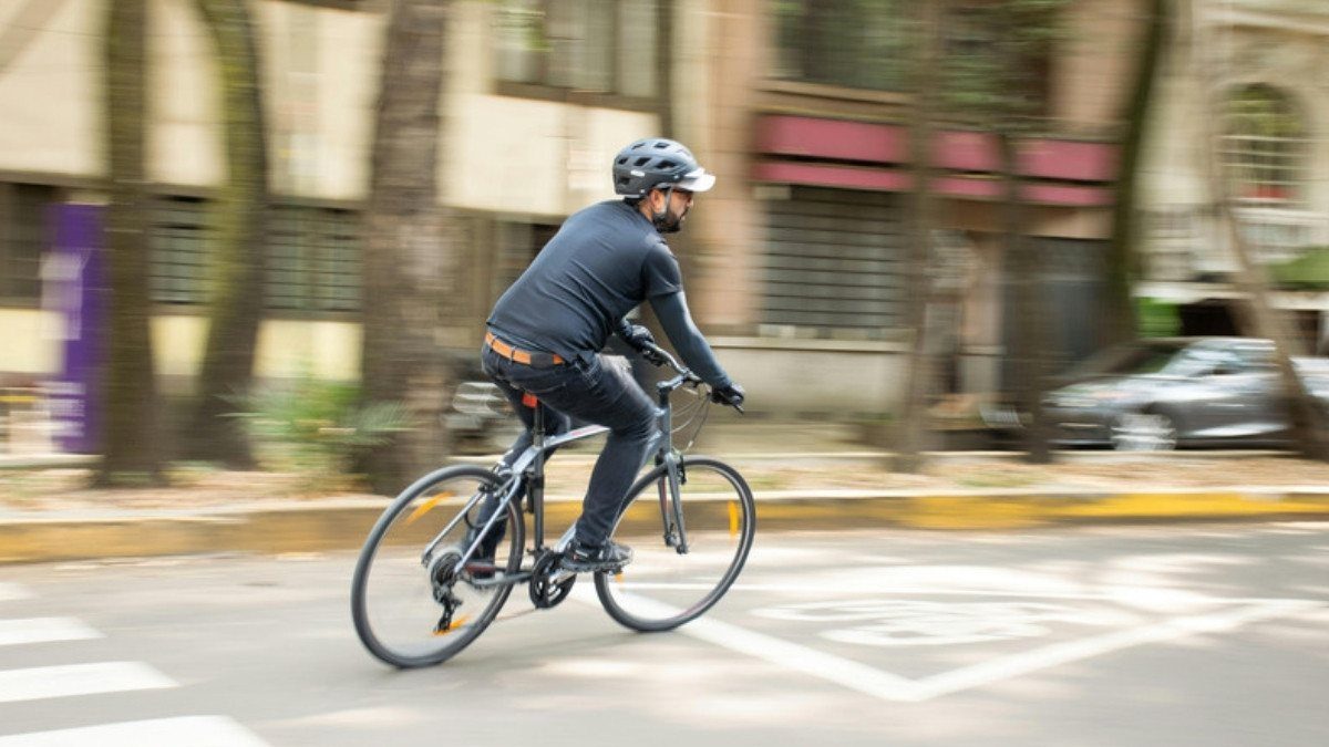 A cyclist in a helmet pedals across a city street as parked cars and buildings blur in the background