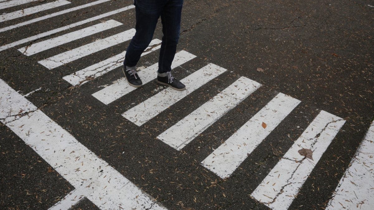 Feet in jeans and sneakers step across the white stripes of a marked crosswalk on worn asphalt