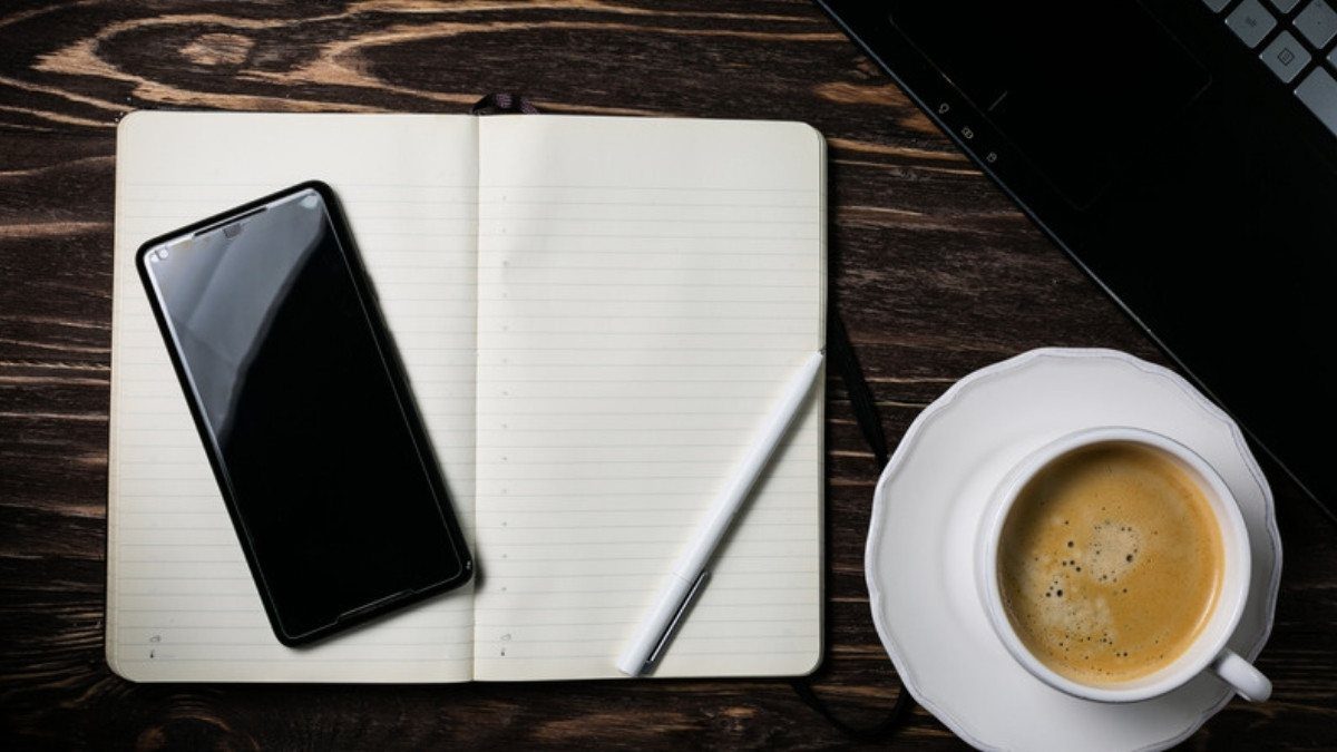An open notebook, a smartphone, a pen and a cup of coffee rest on a dark wooden table before a call
