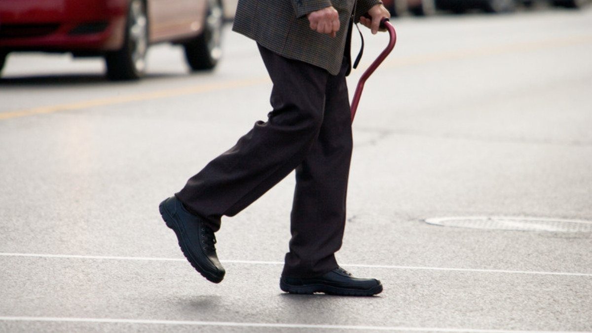 An older man leaning on a cane steps across a city street while cars wait in the lane behind him