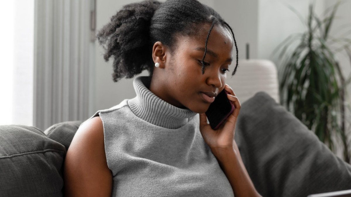 A young woman listens closely on her phone from a gray sofa at home, taking a call at her own pace