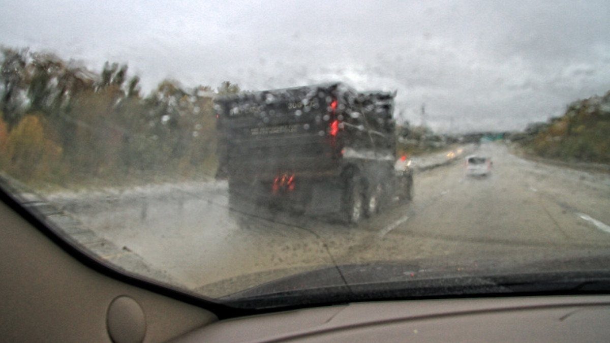 Rain streaks a windshield as a dump truck rolls ahead on a wet highway, conditions that call for slower speeds