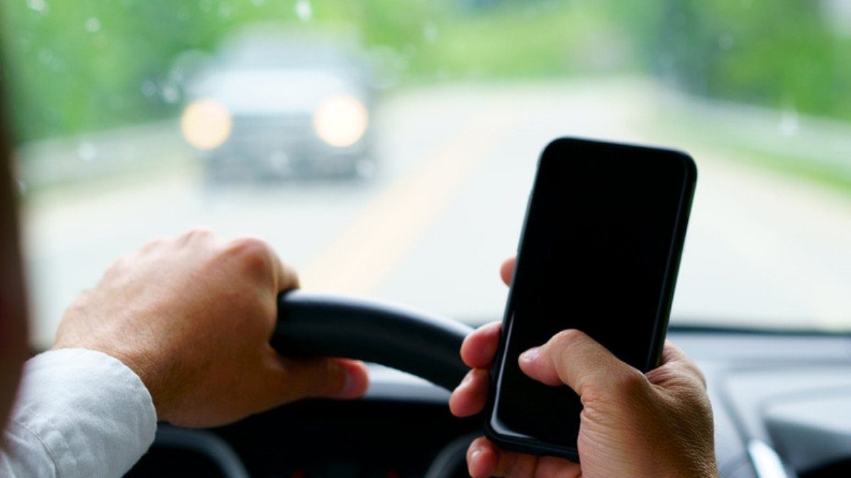 A driver grips the wheel with one hand and holds a phone in the other while an oncoming car approaches on a tree lined road