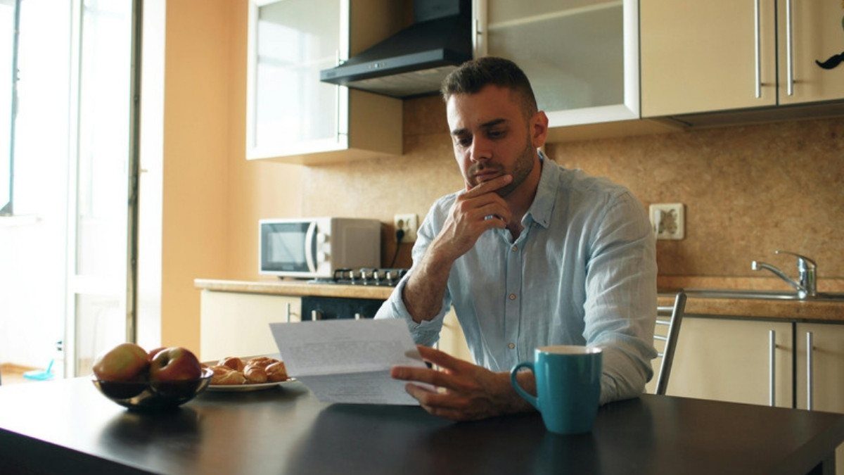 A man reads a letter closely at his kitchen table with a mug of coffee nearby, taking his time before he signs anything