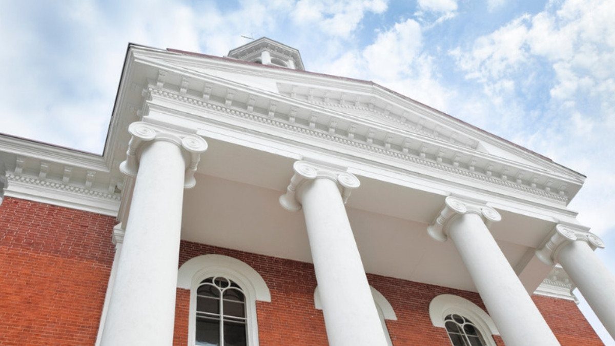 White columns rise in front of a red brick courthouse under a bright sky with scattered clouds