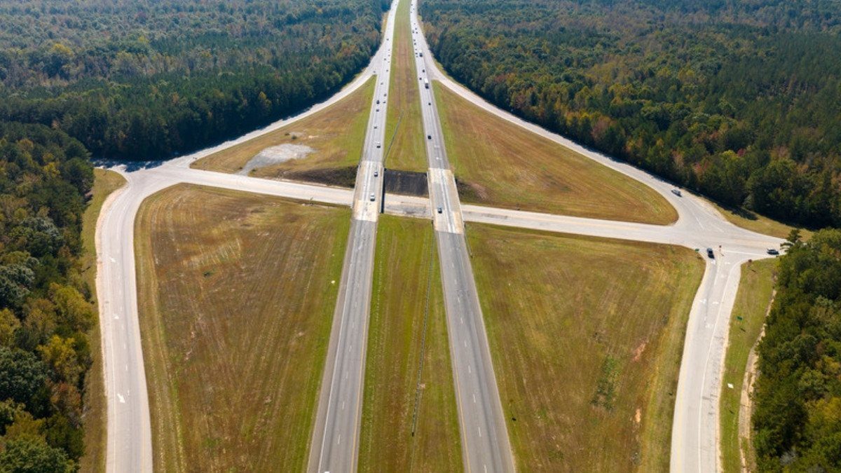 Cars and trucks travel a divided interstate that cuts through thick woods past an overpass and a pair of exit ramps