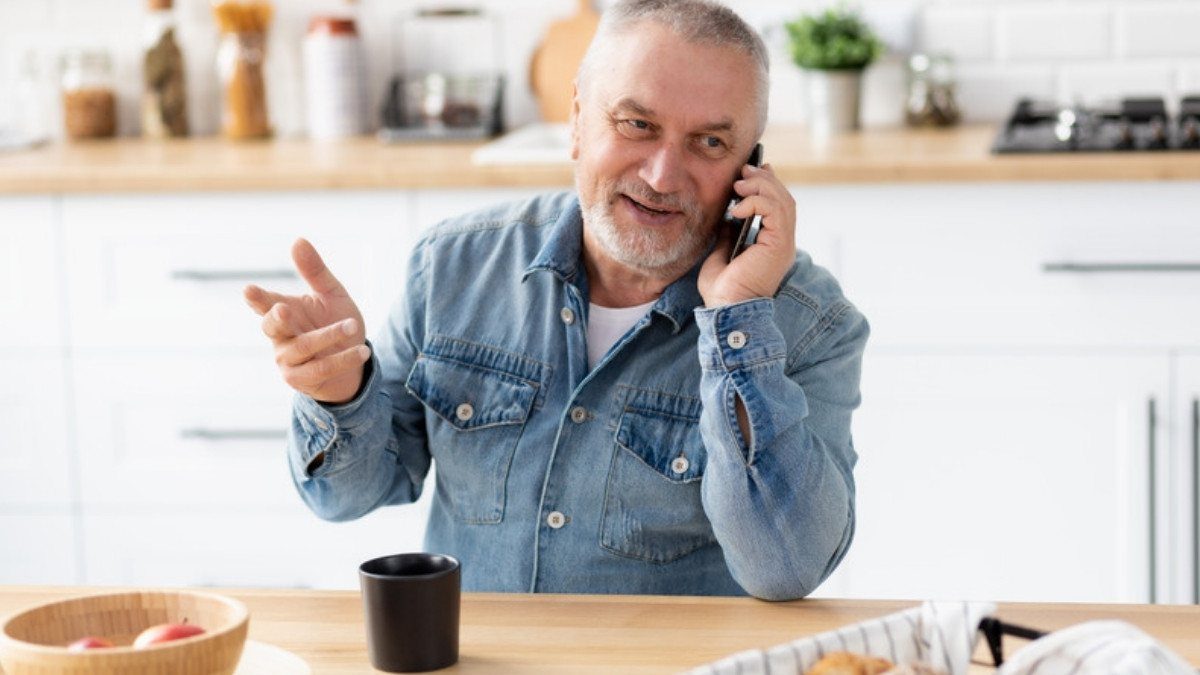 A gray haired man in a denim shirt talks on his cell phone at a sunny kitchen table with a mug and a fruit bowl nearby