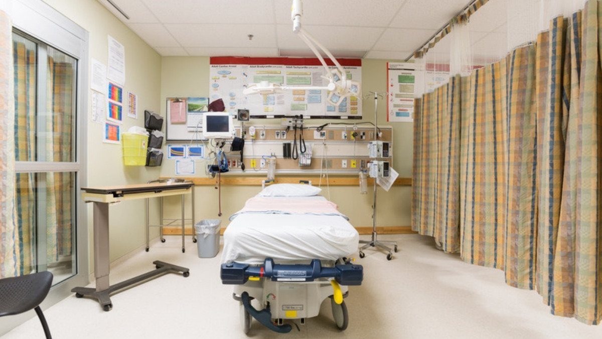 An empty emergency department bay holds a made stretcher, monitors and wall equipment beside a plaid privacy curtain