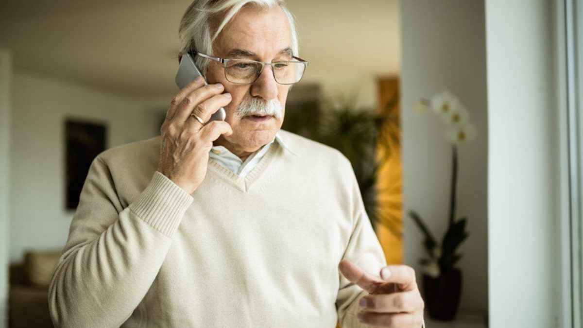 An older man in a cream sweater talks on his phone at home, standing near a bright window with a plant behind him