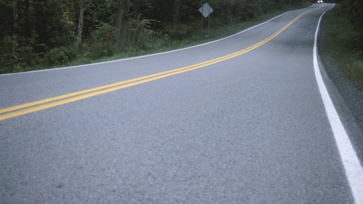 A double yellow center line curves along a wooded two lane road as a car approaches in the distance with its headlights on