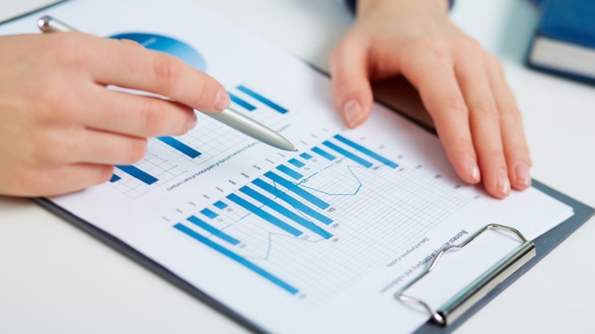A pen points to blue bar charts on a clipboard as two hands review a printed report of figures