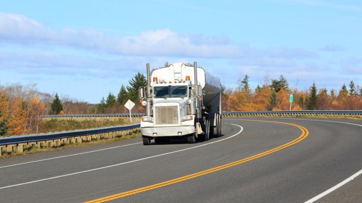A white tanker truck rounds a curve on a two lane highway marked with a double yellow line and lined with autumn trees