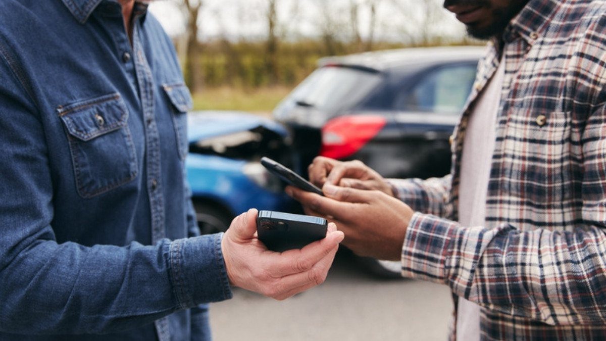 Two drivers hold their phones and trade insurance details while the damaged cars sit behind them on the roadside