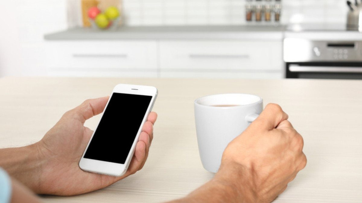 A man holds his phone beside a coffee mug at the kitchen table, taking a moment before calling the insurance company back