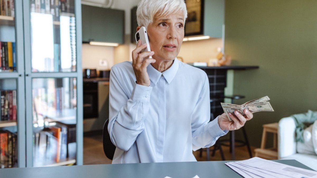 An older woman holds cash and a phone at her kitchen table, weighing a quick offer from the other driver's insurance company