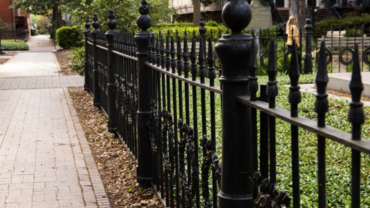 Black wrought iron fencing lines a brick sidewalk in front of a shaded yard on a quiet residential street