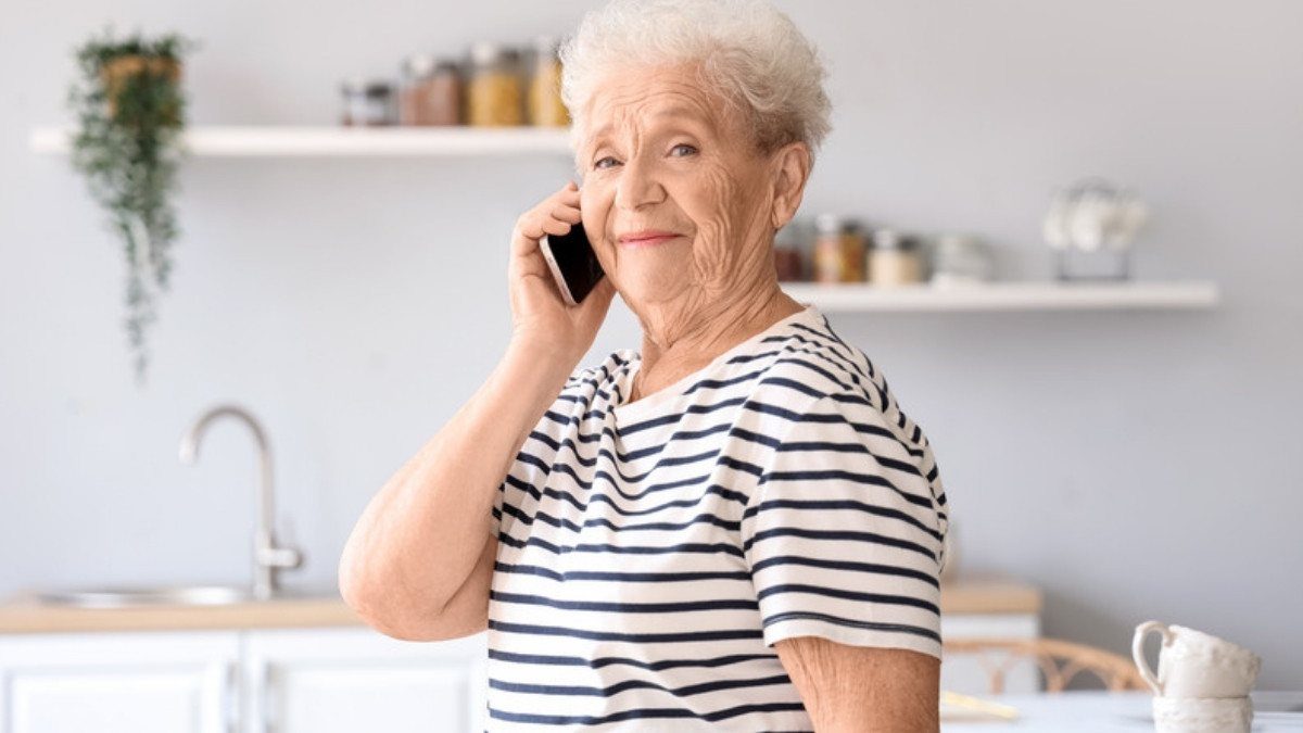 A smiling older woman in a striped shirt talks on her cell phone in a bright kitchen at home