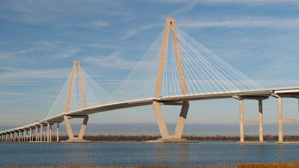 The Arthur Ravenel Jr. Bridge stretches across the water in Charleston under a soft blue sky, with golden marsh grass in front
