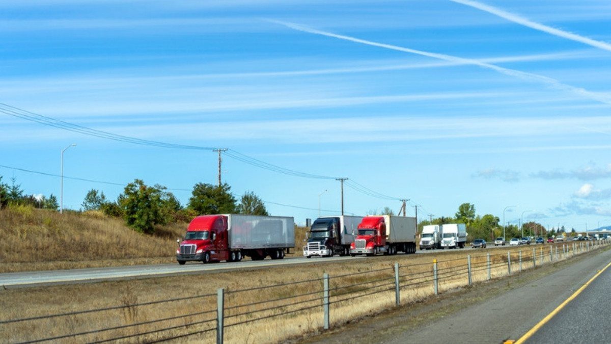A line of tractor trailers and cars travels a divided interstate past dry grass and a wire fence on a sunny afternoon