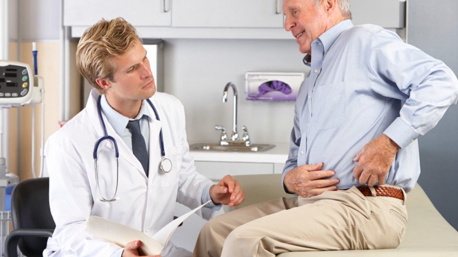 A doctor in a white coat holds a chart while examining an older man who presses a hand to his hip.