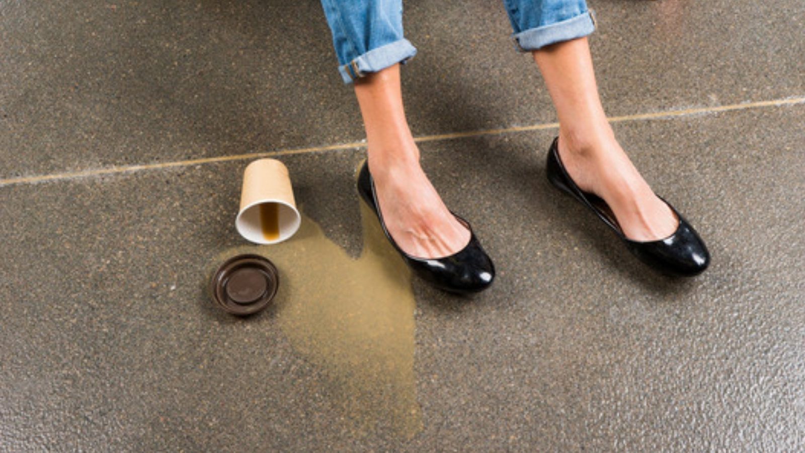 A shopper's feet rest beside an overturned coffee cup and a puddle of spilled coffee on a store floor.