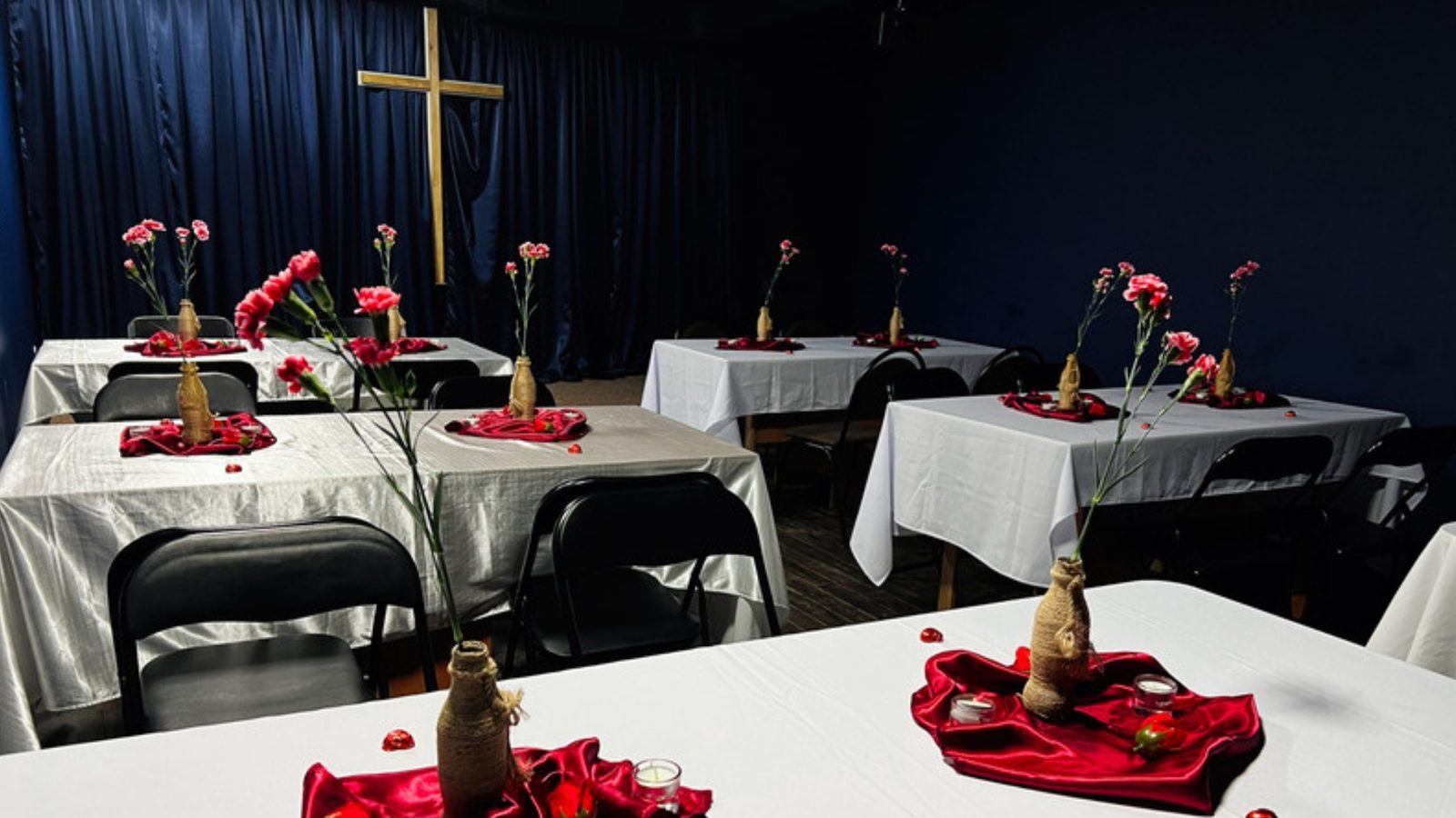 Tables with white cloths, red carnations and candles fill a church fellowship hall beneath a wooden cross.