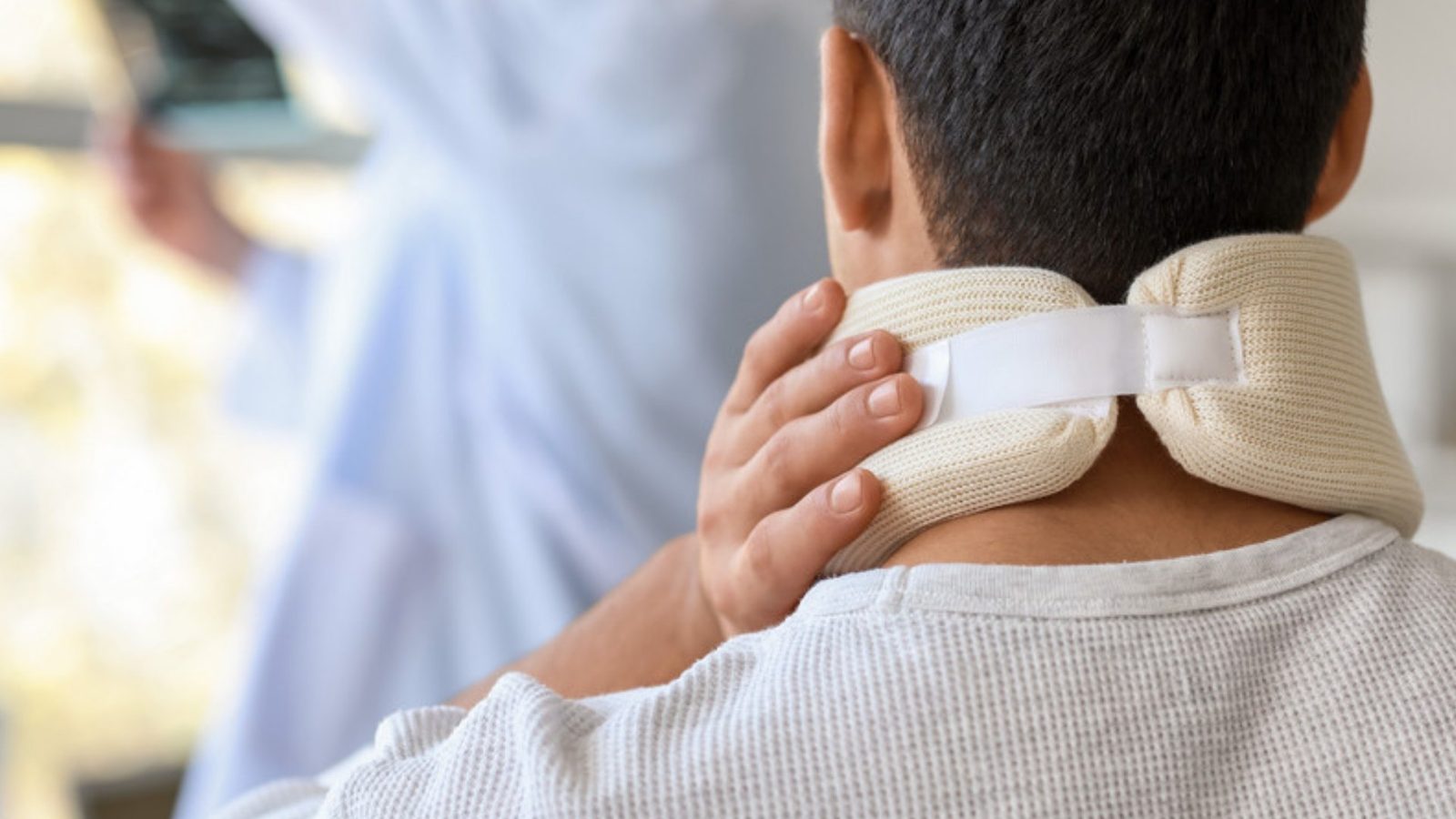 A man wearing a cervical collar holds the back of his neck during a visit to a doctor's office.