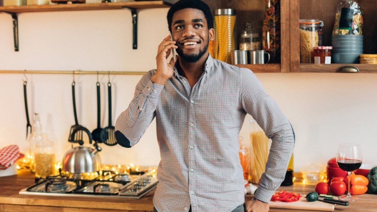 A smiling man in a checked shirt talks on his cell phone while standing in his kitchen near the stove