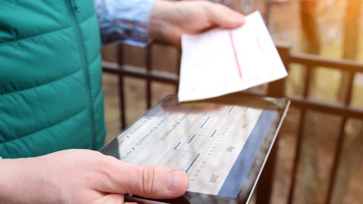 Hands hold a tablet showing a driver log grid while another hand offers a paper slip, the records a carrier keeps