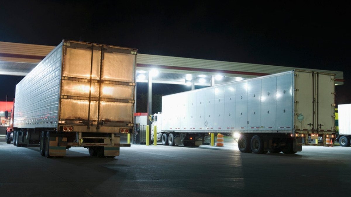 Two tractor trailers sit under the bright canopy of a truck stop fuel island late at night during a long haul