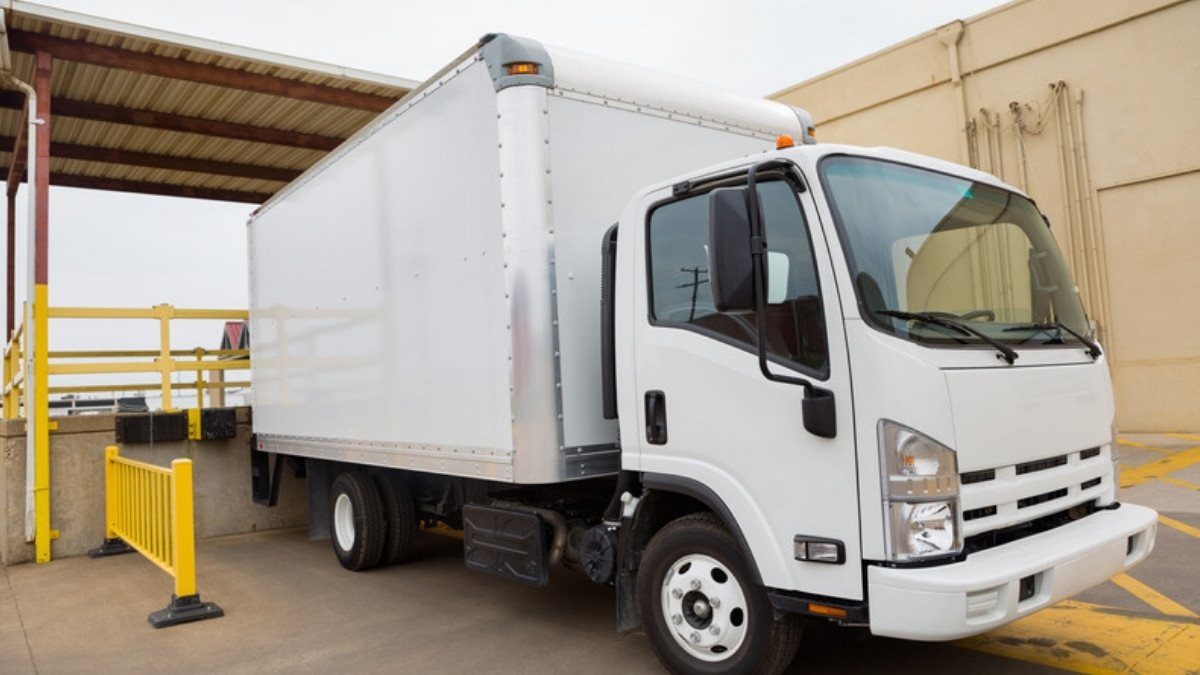 A white box truck waits at a covered warehouse loading dock next to bright yellow safety railings