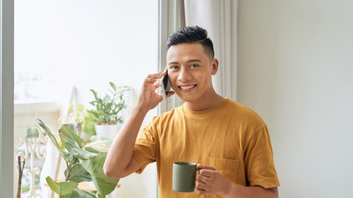 A young man holds a coffee mug and talks on the phone at home, following up after a minor car accident left him sore