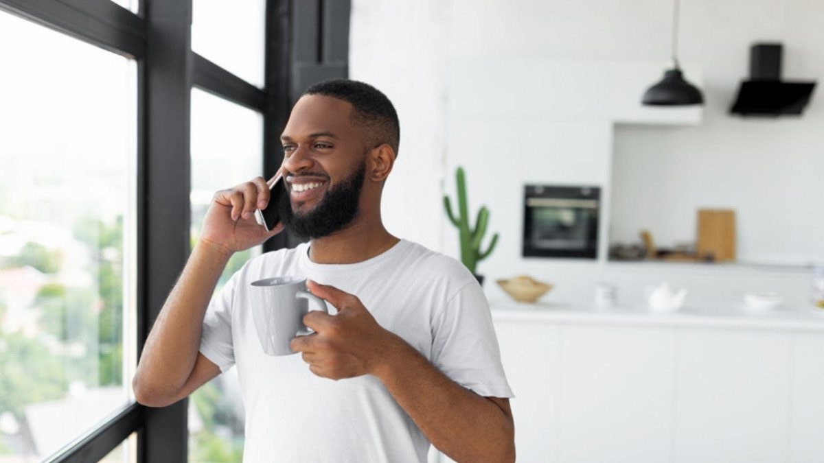 A smiling man talks on the phone with a mug in hand by a sunny window, making a call about a rear end collision