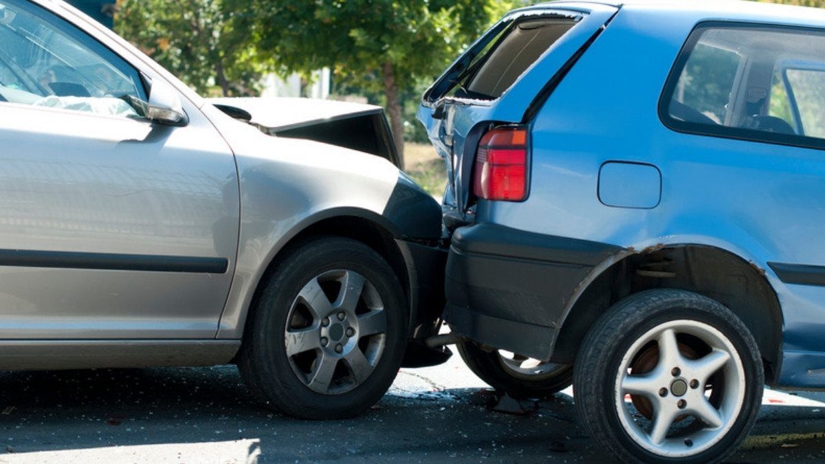 A silver sedan sits wedged into the back of a blue hatchback on a sunny street after a rear end collision