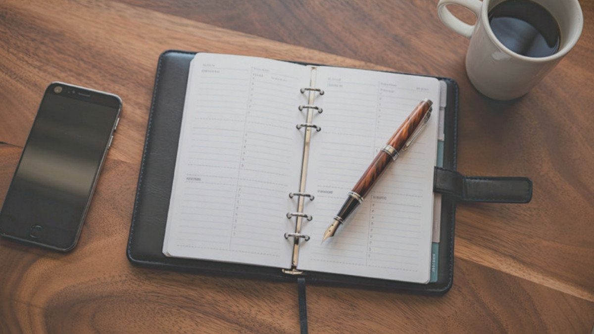 An open planner, a pen, a phone and a cup of coffee sit on a wooden table, ready for listing each name tied to a truck accident