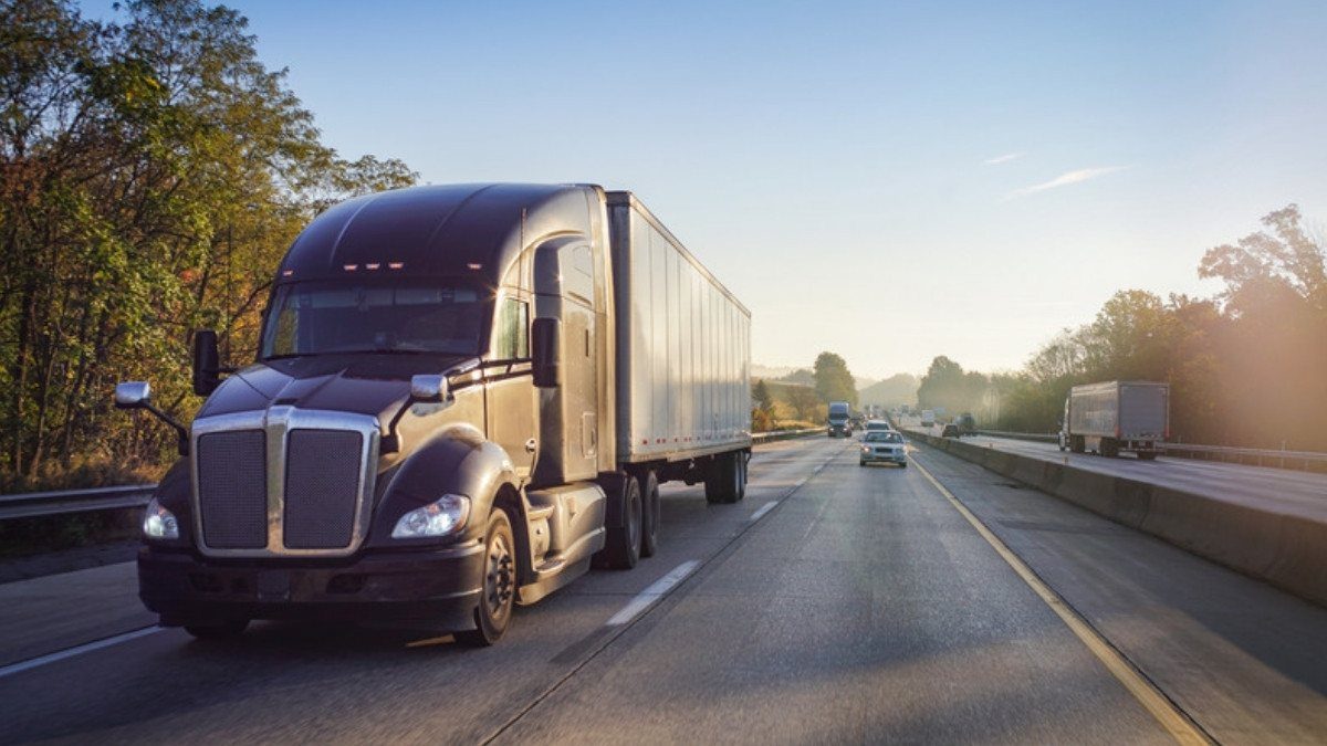 A semi truck pulling a white trailer travels a divided highway lined with trees, where one truck accident can involve many companies