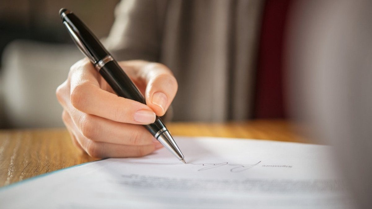 A hand signs a typed document with a black pen at a wooden desk, much like the papers that open an estate in probate court