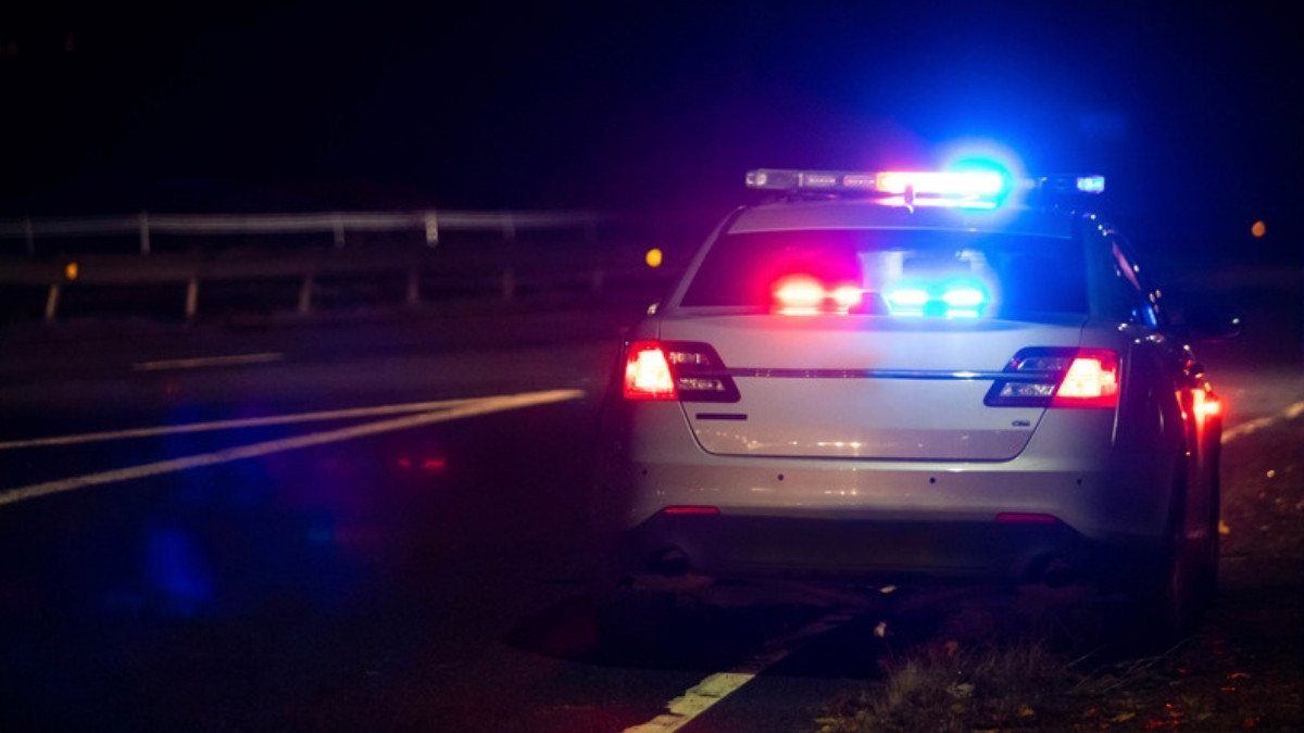 A patrol car with red and blue lights flashing sits on the shoulder of a dark road at night after a call